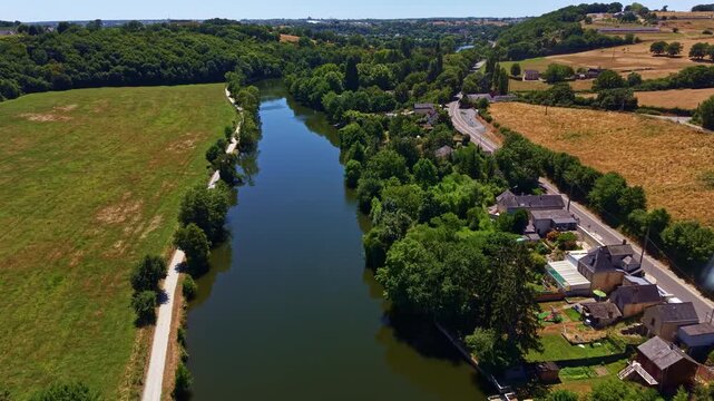 Drone flying above the Mayenne River bordered by rural homes, farmland and forests in western France.