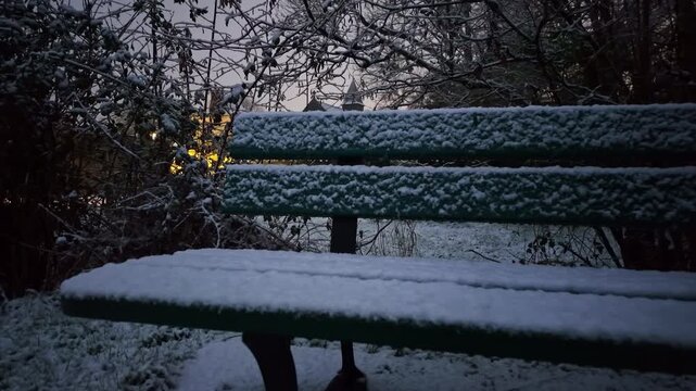 Ground-level push in toward a snow-covered park bench surrounded by frosted trees with a church visible in the background during a peaceful winter evening.