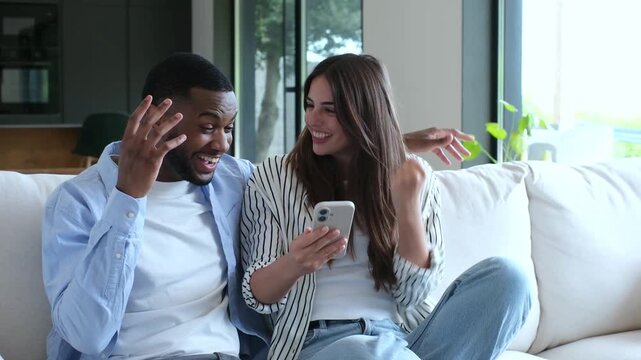 Excited young interracial couple looking at a smartphone, celebrating an online win, getting good news, or achieving a shared goal together while sitting on the sofa in their living room	