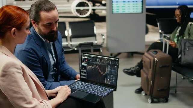 Two business delegates monitor trading data and stock exchange graphs on a laptop in an airport terminal, tracking financial forecasts and trends while waiting near the gate before departure.