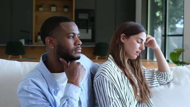 Unhappy young interracial couple sitting on the couch after a fight, avoiding eye contact and conversation, illustrating communication breakdown, marital issues, and family conflict