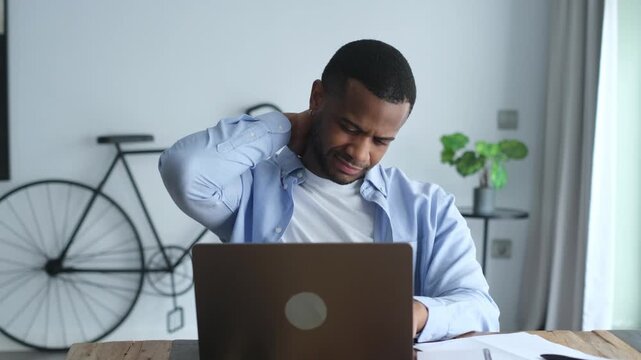 Young overworking african american man feeling unwell, tired and exhausted from long sedentary work on the laptop, massaging his painful neck with his hand and grimacing with discomfort	