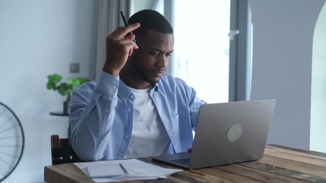 A young Black man concentrates deeply while working on his laptop at a modern home office desk. He thoughtfully reviews information before taking notes, showcasing dedication to his tasks or studies.