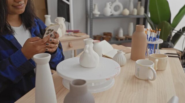 Young woman texting on smartphone beside a white ceramic vase and mugs on a wooden table in studio; calm creativity.