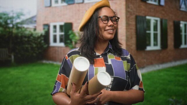 Young african american woman holding rolled blueprints with hands, smiling beside a brick building while wearing a yellow beret; optimism creativity design.