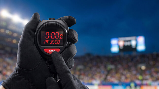 Closeup of a person wearing black gloves holding a digital stopwatch showing 0000 and paused, with a blurred stadium background