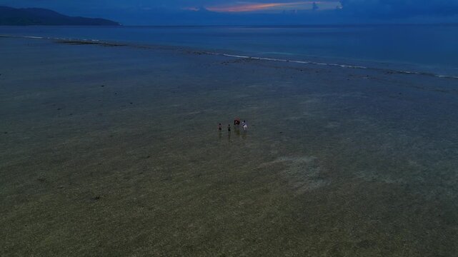 People and horses walking through shallow, clear ocean water under a dramatic sky at twilight during low tide. Majestic aerial view drone shot panorama from above