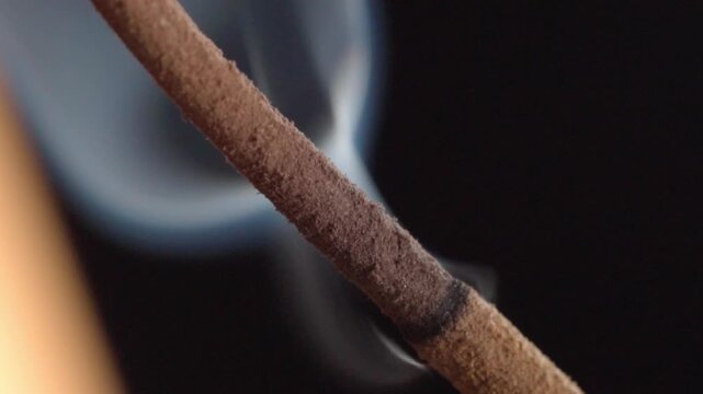 Macro close up of burning incense stick with rising smoke against black background. Detailed view of glowing incense releasing thin white smoke with textured surface