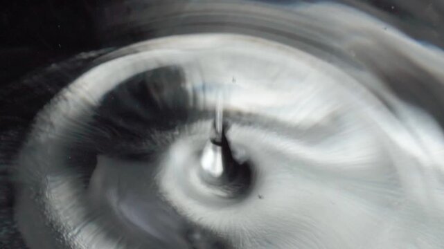 Closeup of liquid droplet striking reflective metallic surface, creating concentric ripples and abstract fluid patterns. Macro view capturing dynamic motion and texture