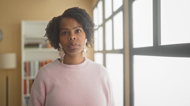 Young woman looks out window in studio, wearing pink sweater and dangling statement earring; contemplation.