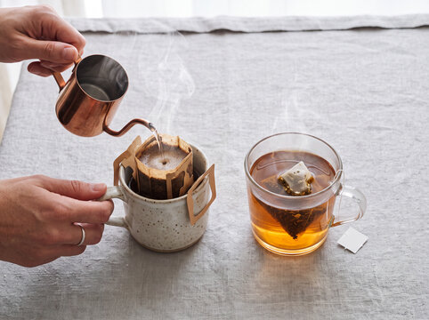 Close-up of hands pouring hot water from a copper pitcher into a drip coffee filter bag over a ceramic mug next to a glass cup of freshly brewed black tea with a tea bag on a linen tablecloth.