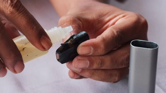 Close-up of an Asian man's hands refilling vape pod with e liquid from dropper bottle, detailed view of pod maintenance and modern electronic cigarette use.