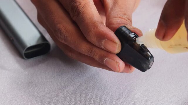 Close-up of an Asian man's hands refilling vape pod with e liquid from dropper bottle, detailed view of pod maintenance and modern electronic cigarette use.