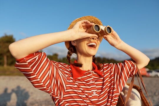 A cheerful woman at the beach uses binoculars, wearing a straw hat and striped shirt. Bright sky and sea behind her; a candid lifestyle moment of outdoor exploration and leisure.