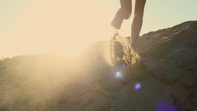 Close-up of barefoot woman running on sandy slope at sunset with sand particles flying toward camera. Freedom, movement, adventure and summer lifestyle concept.