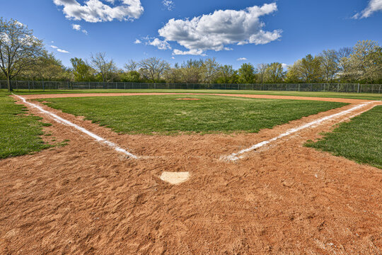 Empty baseball field with home plate in foreground, green grass, trees in background, and partly cloudy blue sky overhead.