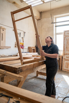 Focused woman woodworker holding antique wooden window frame and examining structure of wood, preparatory work before restoration, female master assessing condition of frame at workplace in workshop.