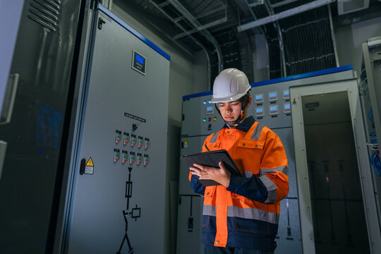 Professional Asian industrial engineer in safety uniform using a digital tablet with stylus to record maintenance data in front of an electrical capacitor bank control panel in a factory.