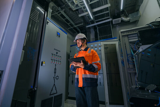 Professional IT network engineer or electrical technician inspecting server rack and industrial power control system using a digital tablet in a modern manufacturing data facility.