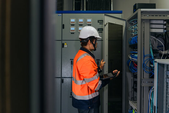 Asian industrial IT technician wearing safety helmet checking network server cabinet and cable connections using a digital tablet for data center infrastructure maintenance.