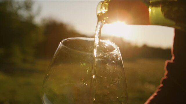 A hand pours a radiant white wine into a glass as sunlight dances through the liquid, capturing the essence of a luxurious evening at a vineyard.