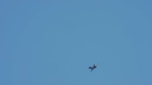 A military fighter jet performs an aerial maneuver, banking and flying across a clear blue sky in slow motion