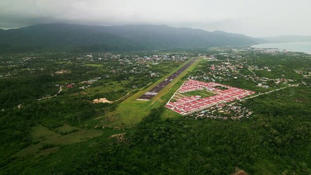 Aerial drone shot of Virac Airport runway surrounded by tropical greenery, subdivision homes, and lush island mountains - Catanduanes, Philippines