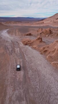 Overhead follow shot tracking dark car driving through eroded desert canyon with textured salt formations and mountain range under cloudy sky. Atacama Desert, Chile