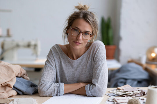 Young woman sits at a sewing table, arms crossed. She wears glasses and a grey sweater, looking calm. Fabric scraps, thread, and a sewing machine lie nearby