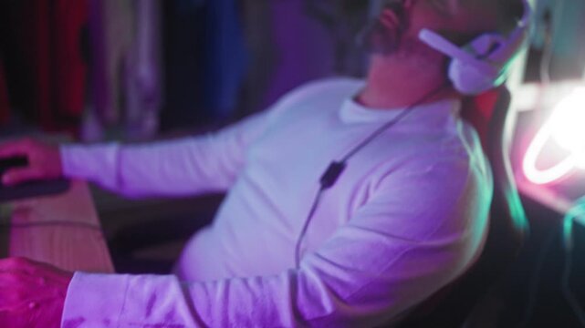 Man in white shirt wearing headset leans back with eyes closed and hand on mouse at keyboard under purple and teal led lights in a gaming studio; exhaustion rest.
