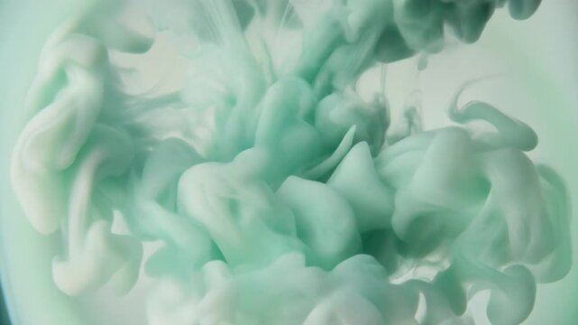 Watercolor pigment blooms through clear water as soft sage green and ivory hues unfurl into organic cloudlike formations under gentle overhead lighting with elegant cinematic macro motion.