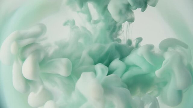 Watercolor pigment blooms through clear water as soft sage green and ivory hues unfurl into organic cloudlike formations under gentle overhead lighting with elegant cinematic macro motion.