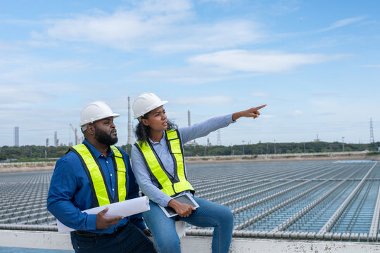 Engineers or industrial workers infrastructure site seated on a railing or a platform solar panel farm environment appears industrial, with several buildings, power lines, and factory structures