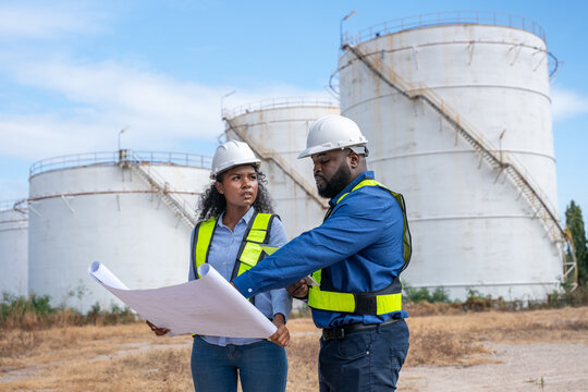 Engineers wearing safety gear, including hard hats examining survey a large blueprint tablet standing industrial facility gas or oil refinery engaged in a job requires high safety standards concept.	