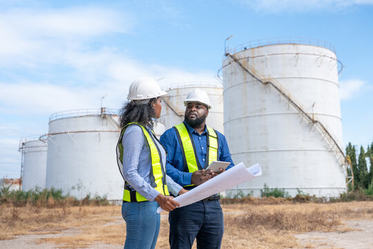Engineers wearing safety gear, including hard hats examining survey a large blueprint tablet standing industrial facility gas or oil refinery engaged in a job requires high safety standards concept.	