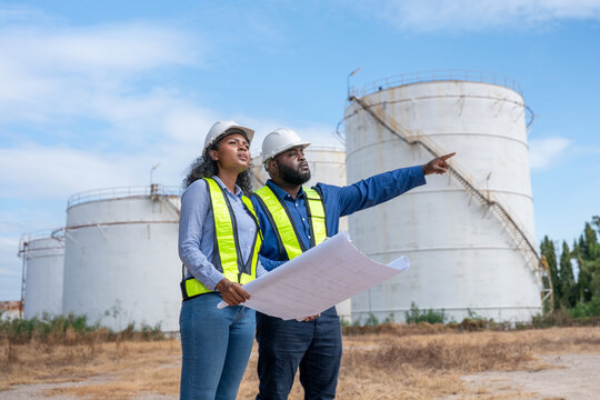Engineers wearing safety gear, including hard hats examining survey a large blueprint tablet standing industrial facility gas or oil refinery engaged in a job requires high safety standards concept.	