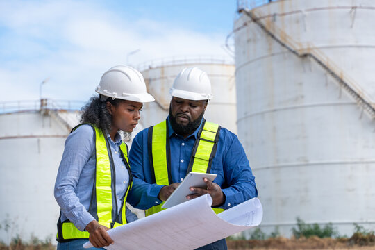 Engineers wearing safety gear, including hard hats examining survey a large blueprint tablet standing industrial facility gas or oil refinery engaged in a job requires high safety standards concept.	