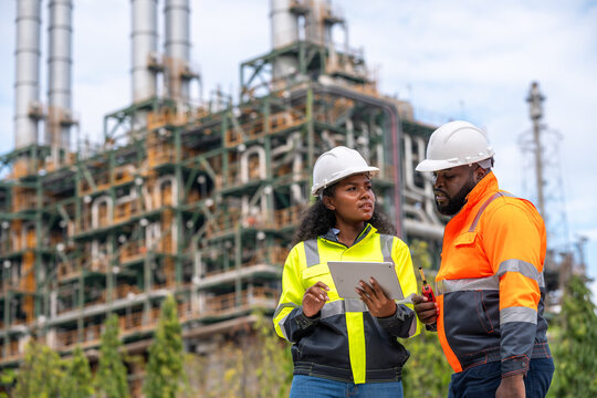 Engineers wearing safety gear, including hard hats examining survey a large blueprint tablet standing industrial facility gas or oil refinery engaged in a job requires high safety standards concept.	