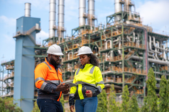 Engineers wearing safety gear, including hard hats examining survey a large blueprint tablet standing industrial facility gas or oil refinery engaged in a job requires high safety standards concept.	