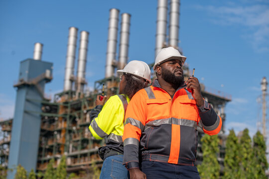 Engineers wearing safety gear, including hard hats examining survey a large blueprint tablet standing industrial facility gas or oil refinery engaged in a job requires high safety standards concept.	