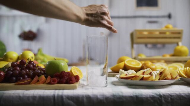 Ice, lemon and orange juice being poured into glass near fruit on kitchen counter - medium shot