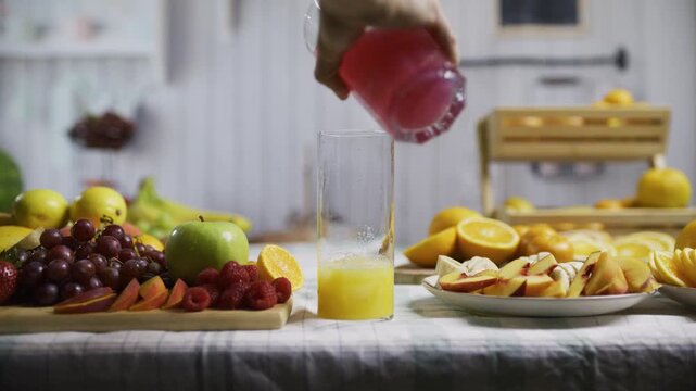Hands of woman dropping fruit and liquids into glass to make drink with surrounding fruit in kitchen - medium shot