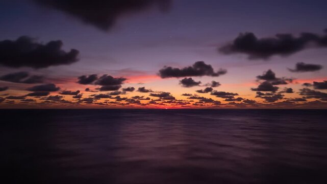 A vibrant orange and deep purple horizon glows across the calm ocean waves as scattered dark clouds drift lazily through the transitional twilight sky while the sun descends below the distant