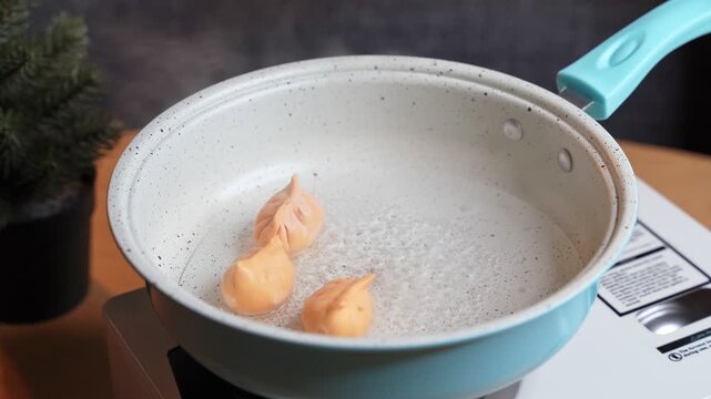 Close up shot of a person's hand placing a fresh orange dumpling into a light blue pan of boiling water. The asian dim sum cooks on a white portable stove on a wooden table indoors.