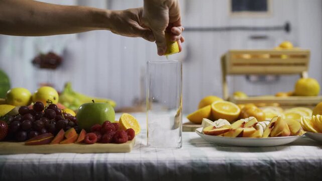 Woman's hand dropping ice, lemon and other juices into glass with surrounding fresh fruit on kitchen counter - medium shot