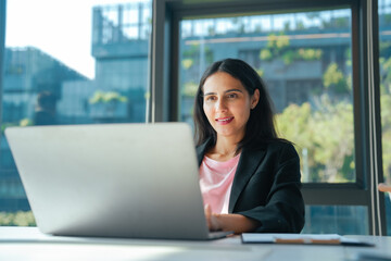 Fototapeta premium Businesswoman working on laptop in office with city view through window. Office workspace for online communication, project planning, remote work and business management tasks.