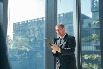 Fototapeta premium Businessman reading tablet in modern office with large window. Professional reviewing business data, planning work, managing projects and using digital technology in workplace.