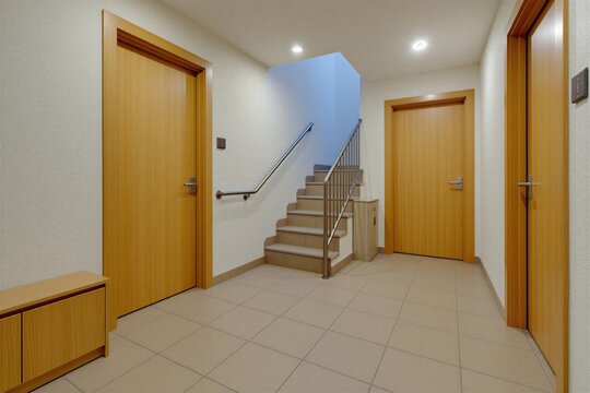 Minimalist hallway with beige tiled floor, light wood doors and white textured walls. Features a short metal-railed staircase and a small utility cabinet. Clean neutral interior passage.