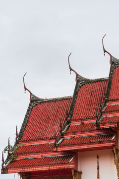Architectural detail of a traditional Thai temple roof with red tiles and Chofah against overcast sky.