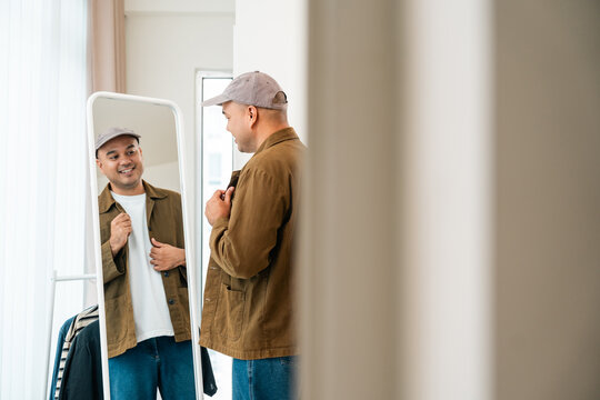 An Asian man looks in the mirror at his attire. He chooses clothes to wear out, matching shirts and pants skillfully. He selects clothes that boost his confidence.